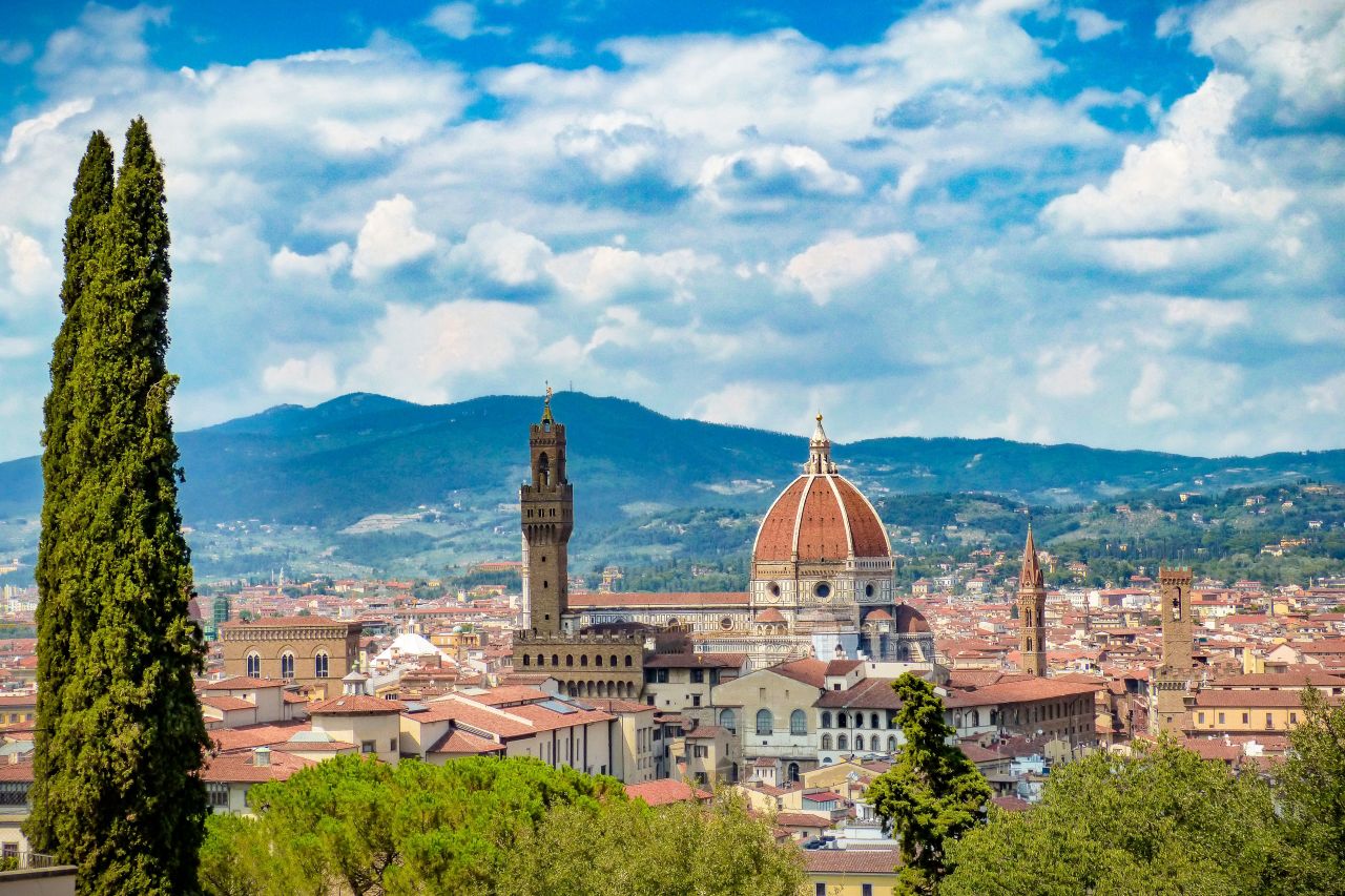 Panoramic view of Florence with the Duomo, Palazzo Vecchio, and cypress trees under a cloudy blue sky.