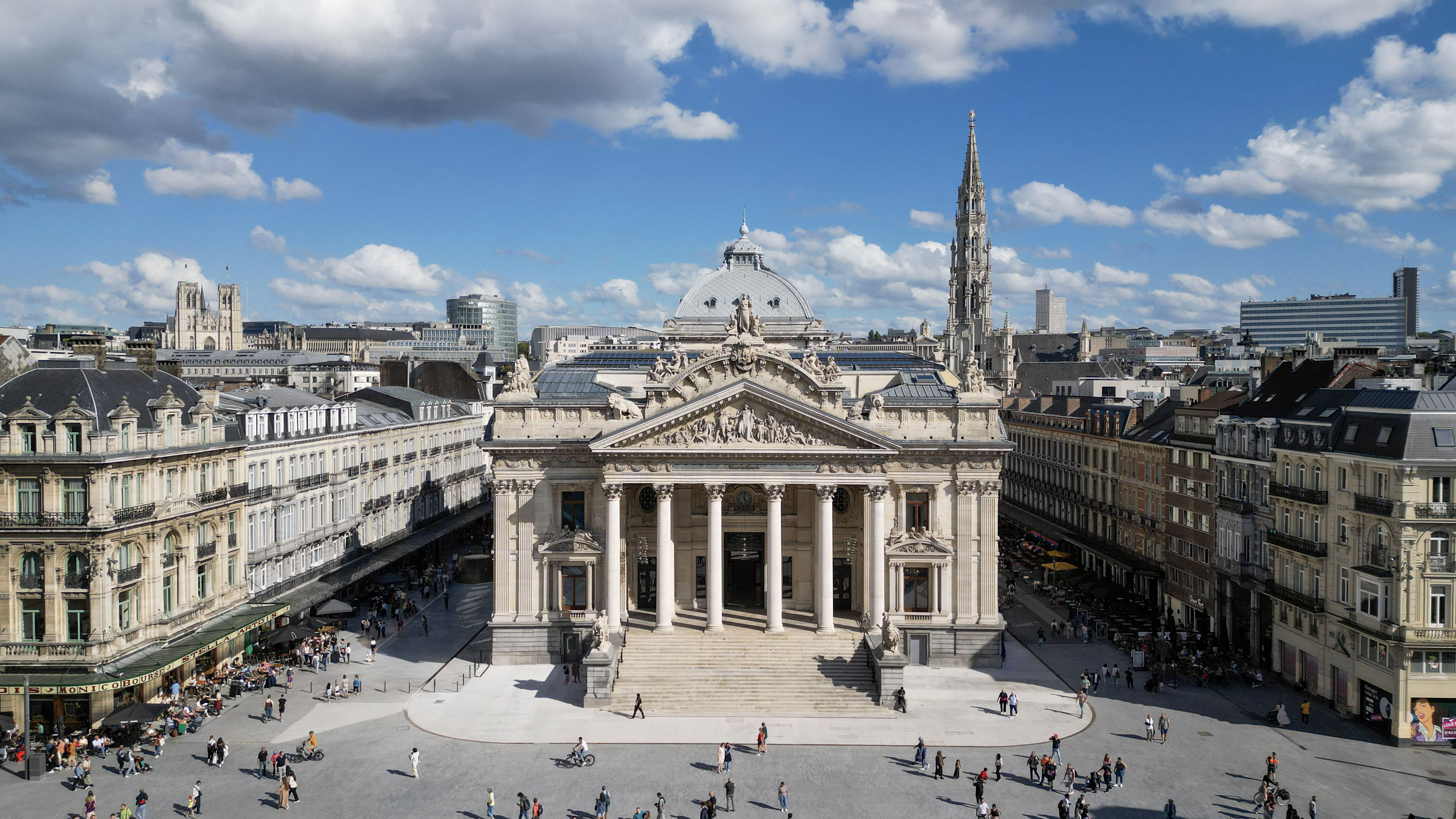 A high-angle aerial view of the historic Brussels Stock Exchange building, surrounded by a lively pedestrian square under a blue sky with white clouds.