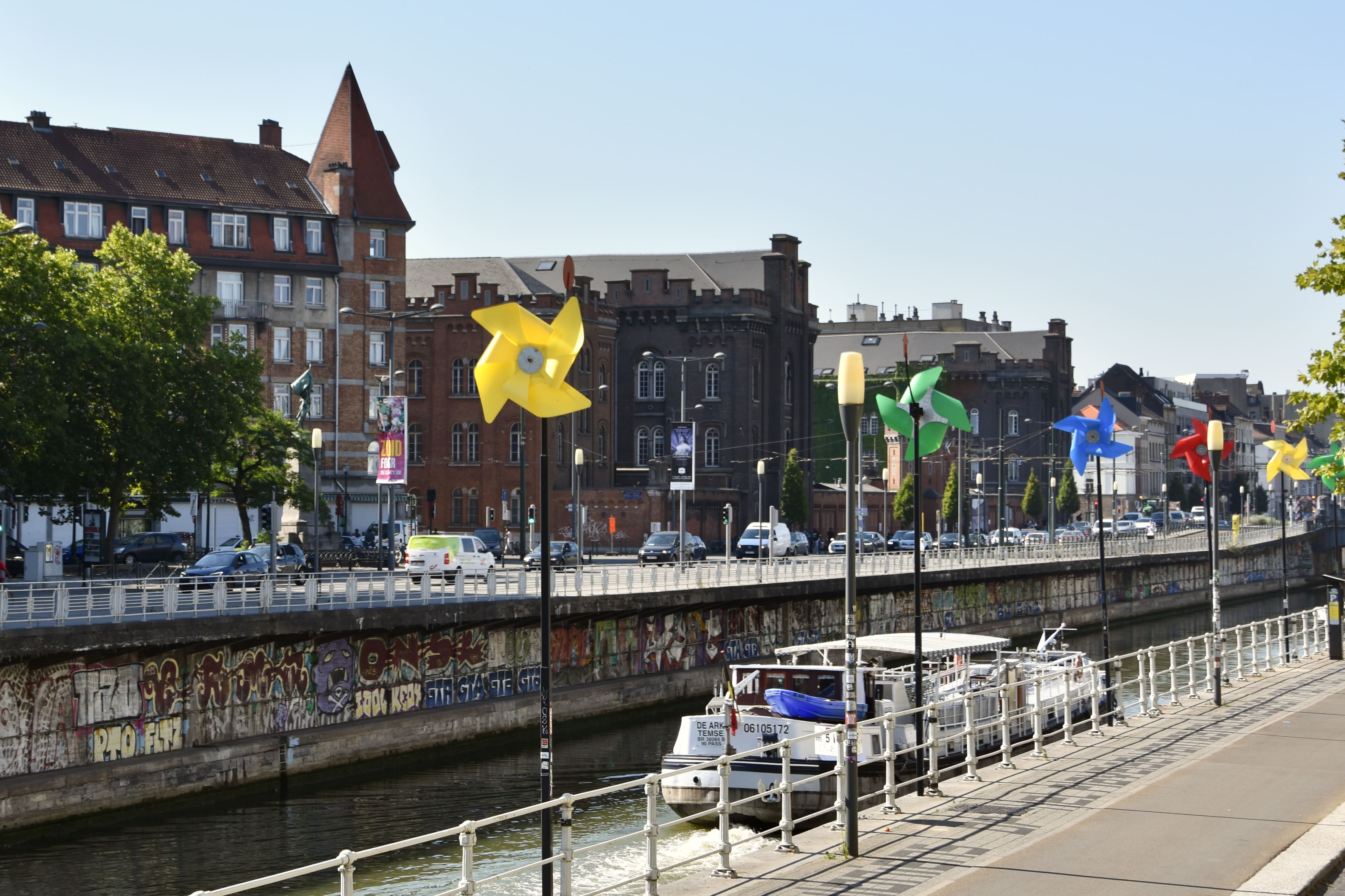 Molinillos de viento de colores (amarillo, verde, azul, rojo) alineados a lo largo del canal de Bruselas, con una barcaza navegando por el agua y edificios urbanos al fondo.
