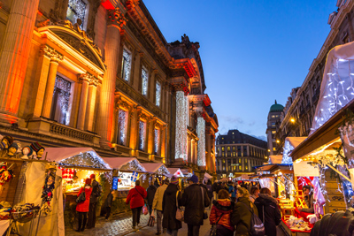 Illuminated chalets at the Brussels Christmas market (Plaisirs d'Hiver) set up in front of the Stock Exchange building lit in warm colors, with passersby strolling at dusk.