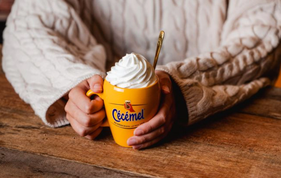 Een persoon in een witte gebreide trui houdt een gele Cécémel-mok vast, gevuld met warme chocolademelk en afgetopt met een flinke toef slagroom, op een houten tafel.