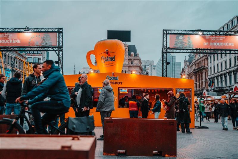 Een grote oranje Cécémel-stand met een gigantische opblaasbare mok op de kerstmarkt van Brussel (Winterpret) op het De Brouckèreplein, met voorbijgangers en een fietser op de voorgrond.