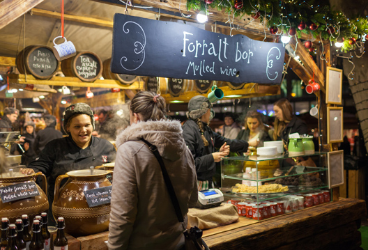 An illuminated wooden chalet at the Brussels Christmas market, displaying a "Forralt bor / Mulled wine" sign, where vendors serve steaming mulled wine to customers in a festive winter atmosphere.