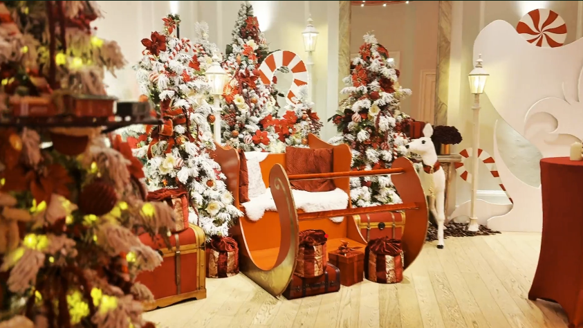 Een magisch kerstdecor binnen in Brussel met een grote rode en gouden slee in het midden, omringd door versierde besneeuwde kerstbomen, cadeaukisten, reusachtige zuurstokken en een beeld van een rendier.