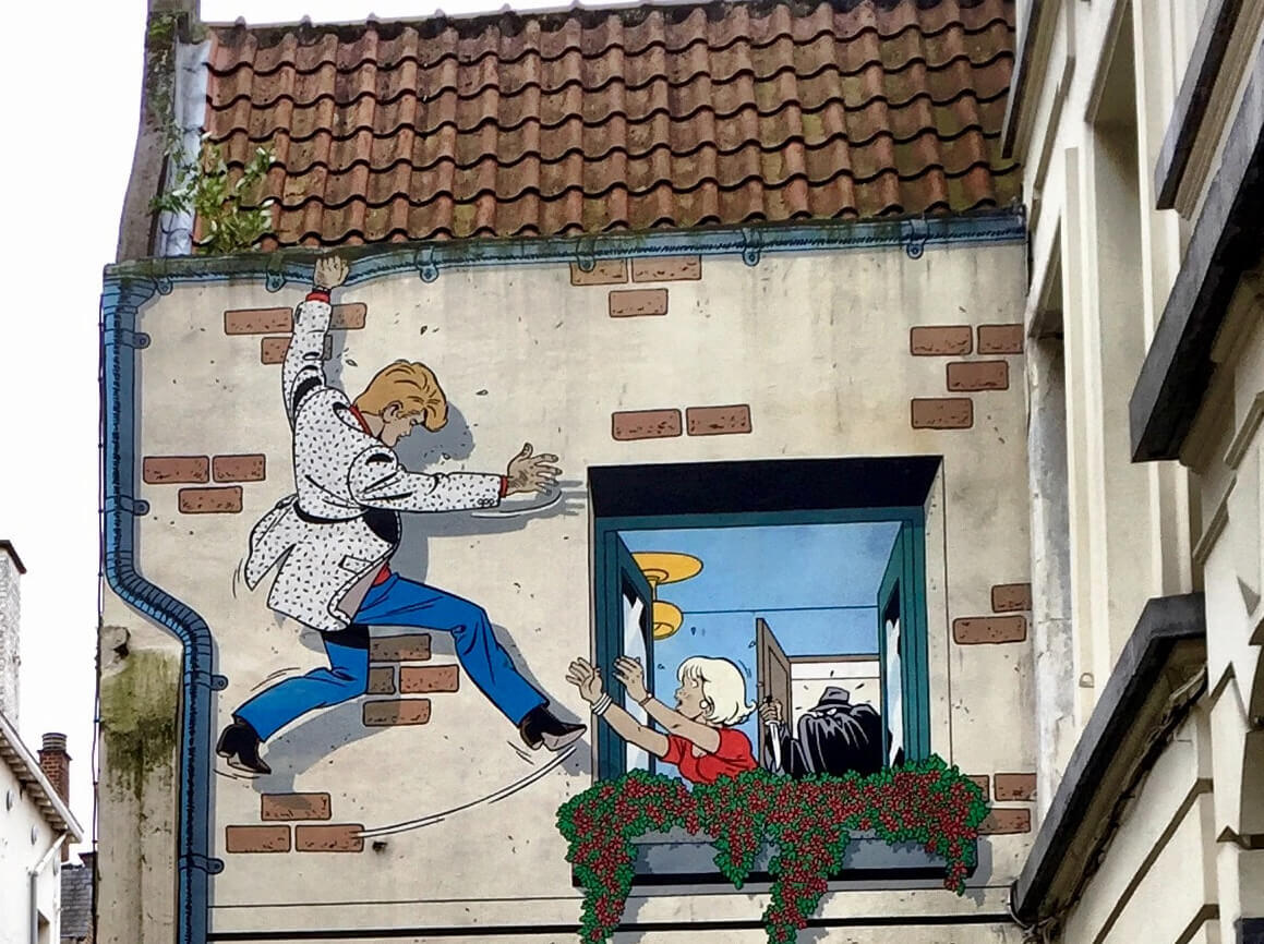 Een stripmuur van de Brusselse Striproute die het personage Ric Hochet voorstelt terwijl hij aan de gevel van een gebouw hangt en probeert een vrouw bij een raam te bereiken.