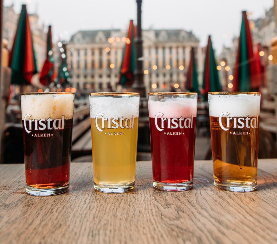 Vier glazen met verschillende soorten Belgisch bier (donker, blond, fruit, amber) van het merk Cristal Alken, op een rij op een houten tafel met de Grote Markt van Brussel op de wazige achtergrond.