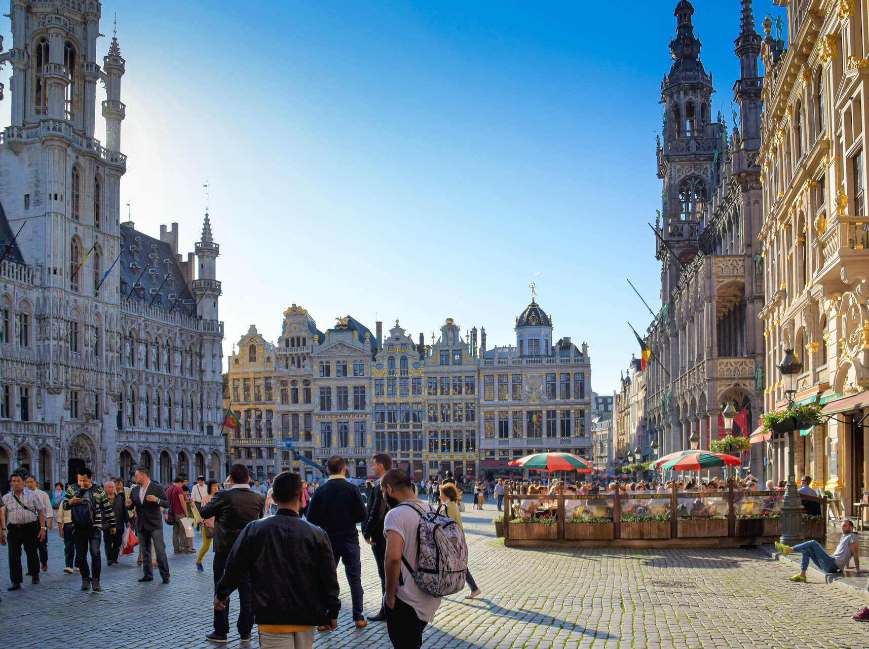 Passersby and outdoor café seating in the famous Grand-Place in Brussels surrounded by magnificent Gothic and Baroque guildhouses under a clear blue sky.