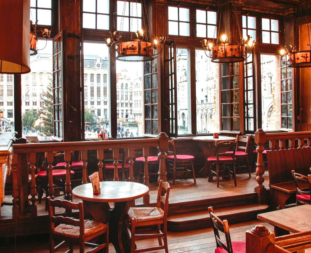 Het warme interieur van een traditionele houten taverne in Brussel, met ronde tafels en grote kroonluchters, dat door de open ramen een adembenemend uitzicht biedt op de historische gebouwen van de Grote Markt.