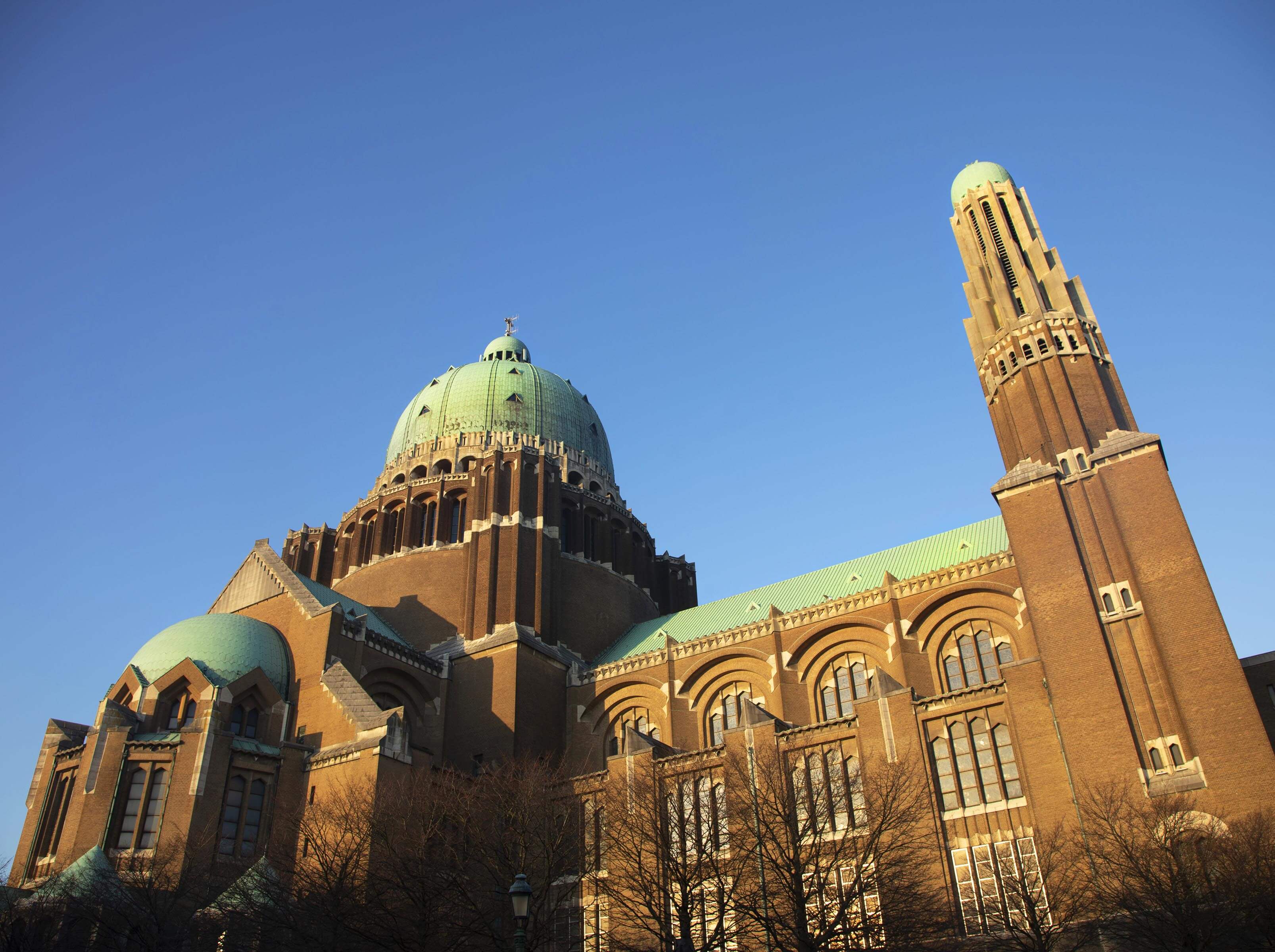 Kikvorsperspectief van de majestueuze Nationale Basiliek van het Heilig Hart te Koekelberg in Brussel, met de nadruk op de grote groene koepel en bakstenen torens onder een blauwe lucht.