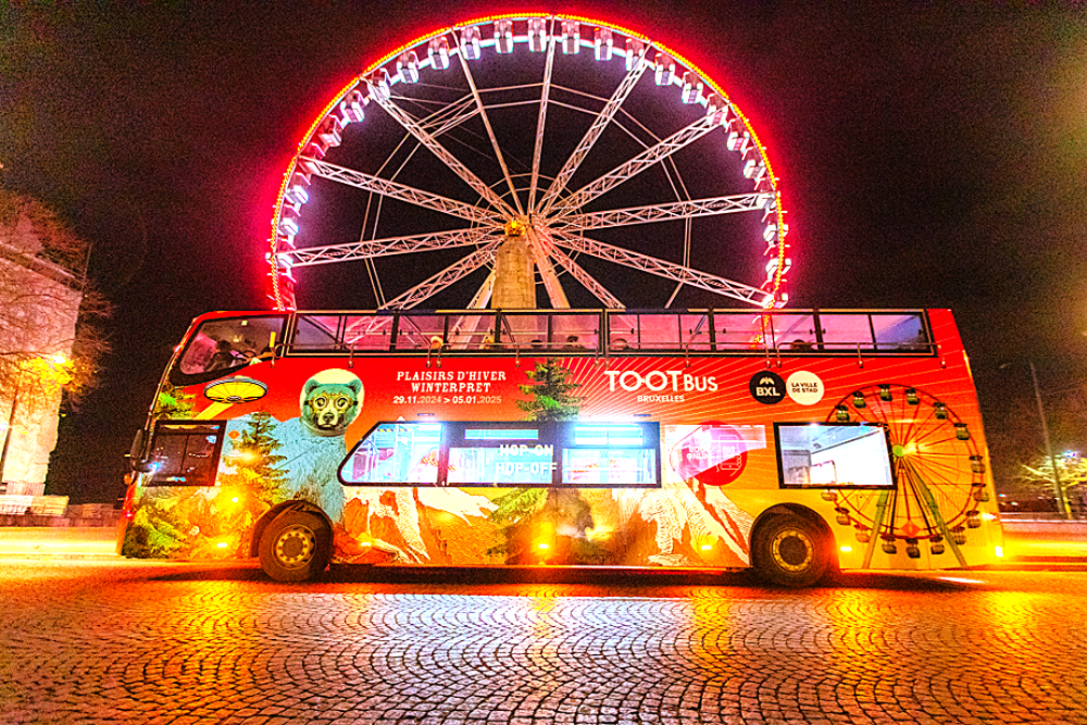 A Tootbus Brussels double-decker bus wrapped in the Winter Wonders (Plaisirs d'Hiver / Winterpret) event branding, parked at night in front of the giant Ferris wheel illuminated in red at Place Poelaert.