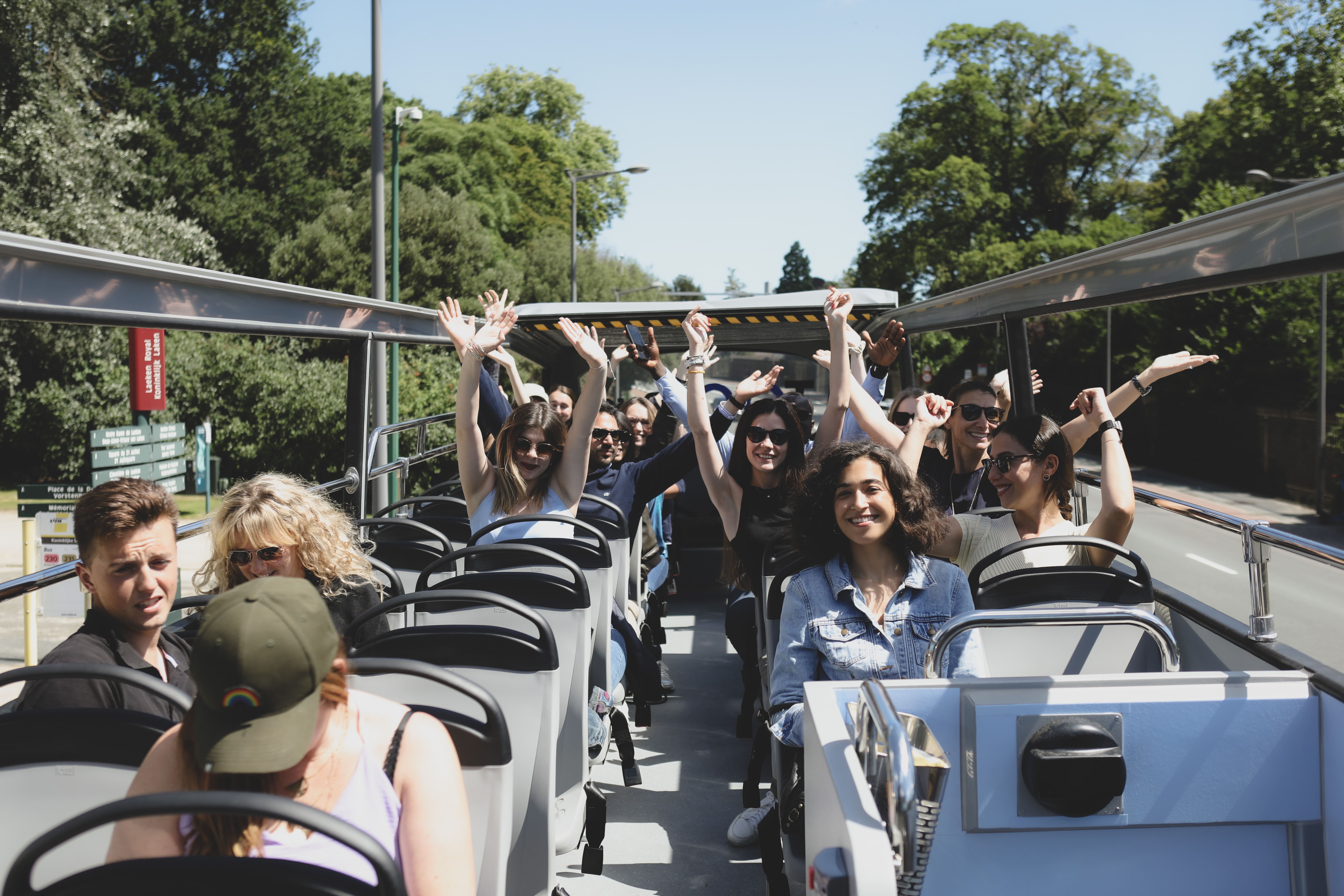 Un grupo de pasajeros entusiastas y sonrientes levanta los brazos a bordo del piso superior abierto de un autobús Tootbus Brussels, disfrutando de un día soleado en la ciudad.