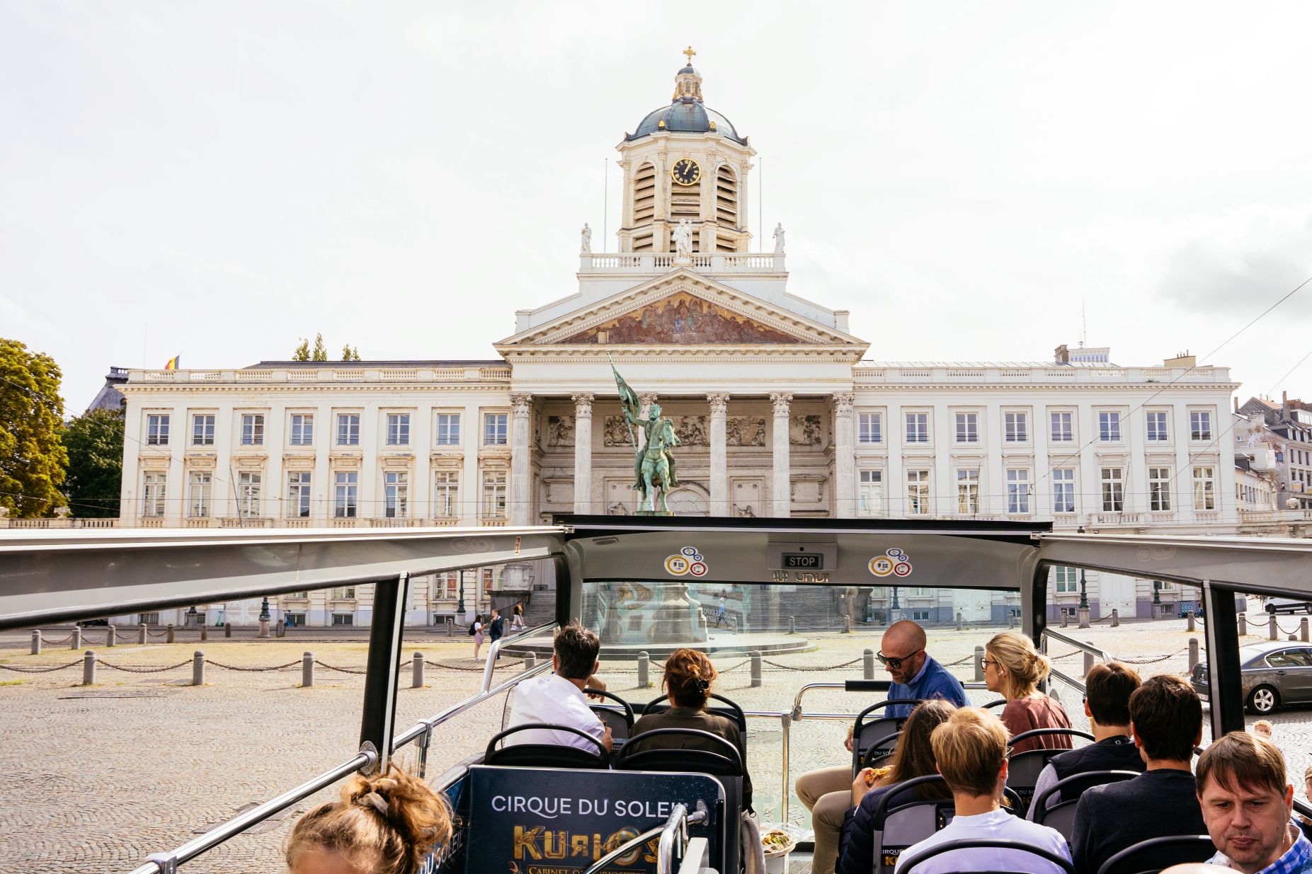 Passagiers op het open bovendek van een Tootbus Brussels die aankomen op het Koningsplein, met de Sint-Jacob-op-Coudenbergkerk en het standbeeld van Godfried van Bouillon op de achtergrond.