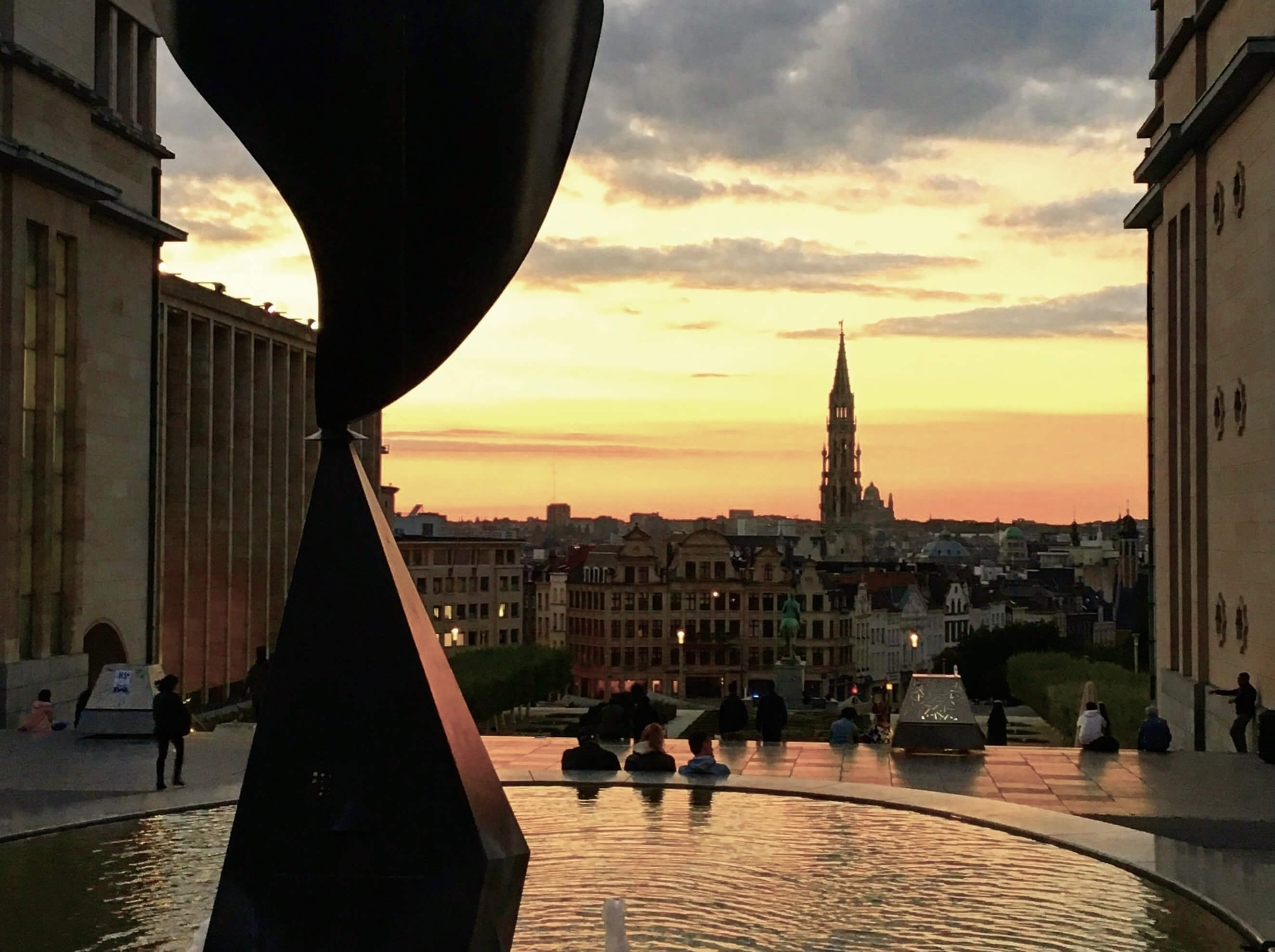 Vue panoramique depuis le Mont des Arts à Bruxelles au coucher du soleil, avec une fontaine au premier plan et la flèche de l'Hôtel de Ville à l'horizon lors d'un tour privé.