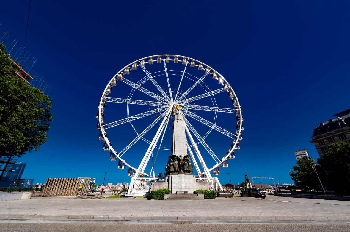 Front view of a large white Ferris wheel on Poelaert Square in Brussels, framing a historic stone monument under a deep blue sky.