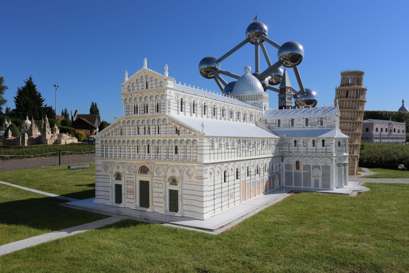 Vue des monuments miniatures de la cathédrale de Pise et de sa tour penchée au parc Mini-Europe à Bruxelles, avec la structure monumentale de l'Atomium en arrière-plan sous un ciel bleu.