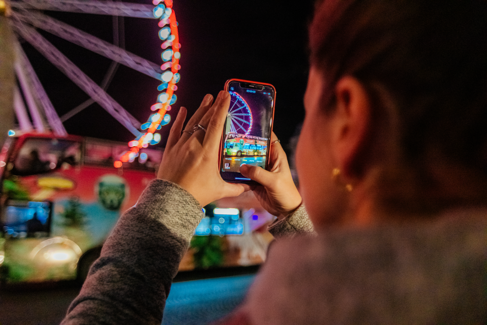Blik over de schouder van een persoon die met hun smartphone een foto maakt van het verlichte reuzenrad en de feestelijke kerstbus van Tootbus, 's nachts in Brussel.