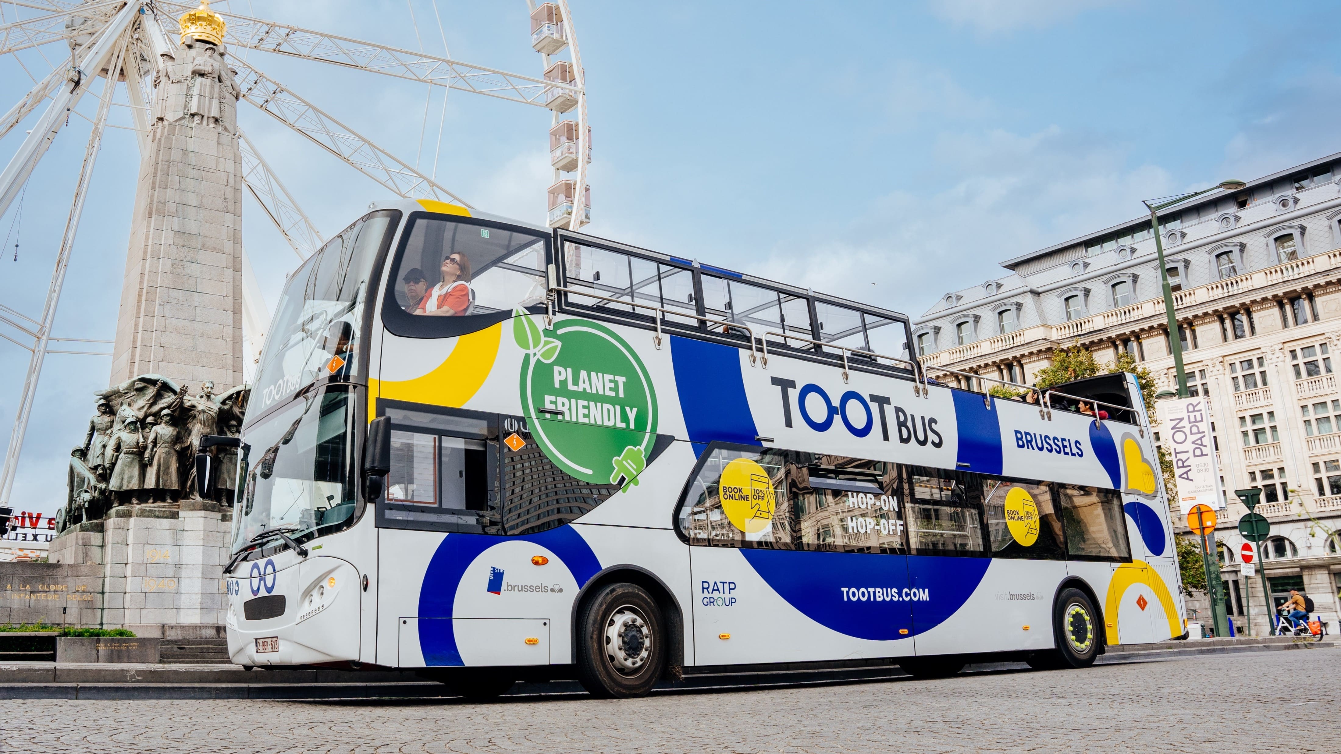 Planet-friendly Tootbus Brussels double-decker tourist bus parked in the city in front of a Ferris wheel and a historic monument under a cloudy sky.