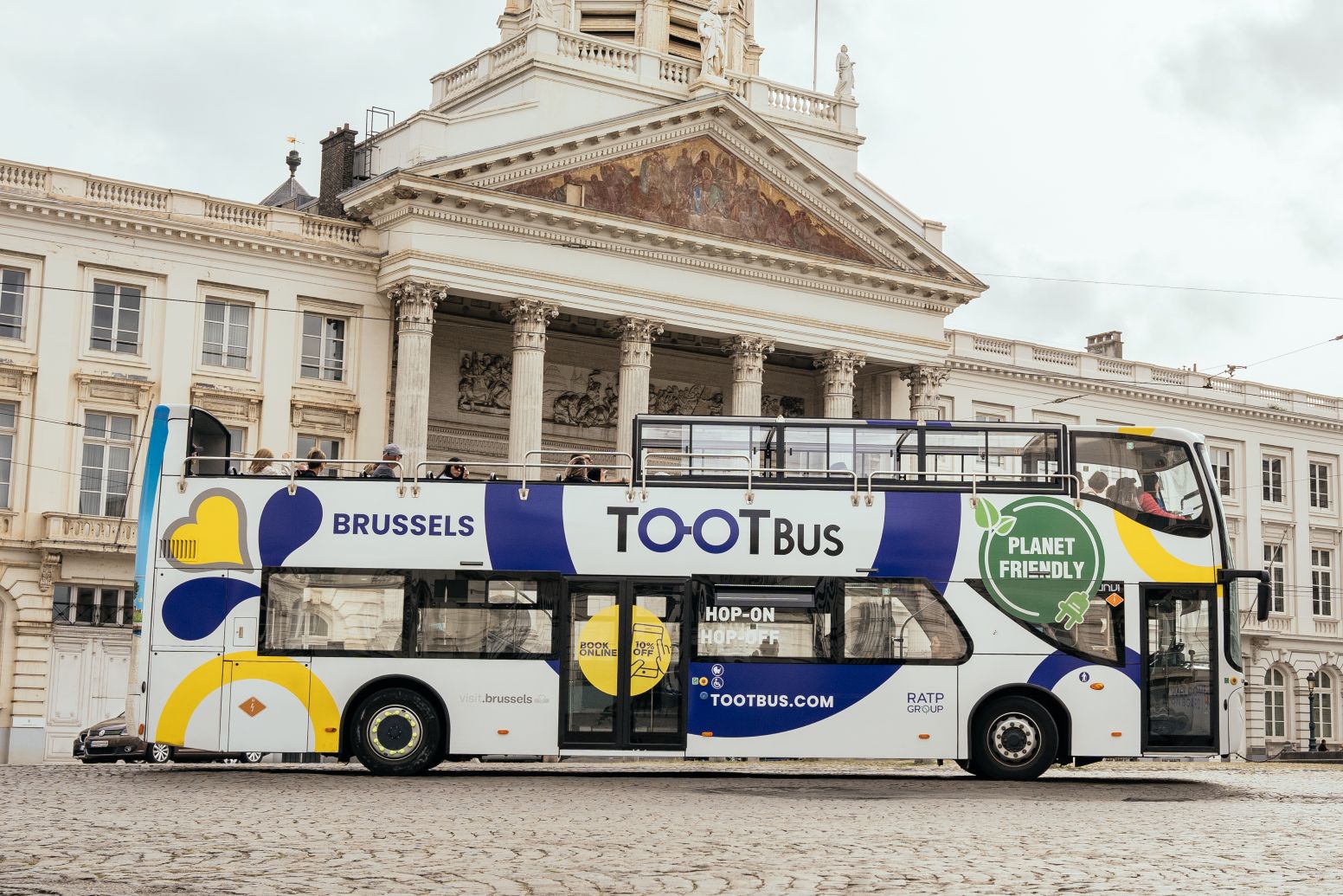 Passagiers op het open bovendek van een Tootbus Brussels die aankomen op het Koningsplein, met de Sint-Jacob-op-Coudenbergkerk en het standbeeld van Godfried van Bouillon op de achtergrond.