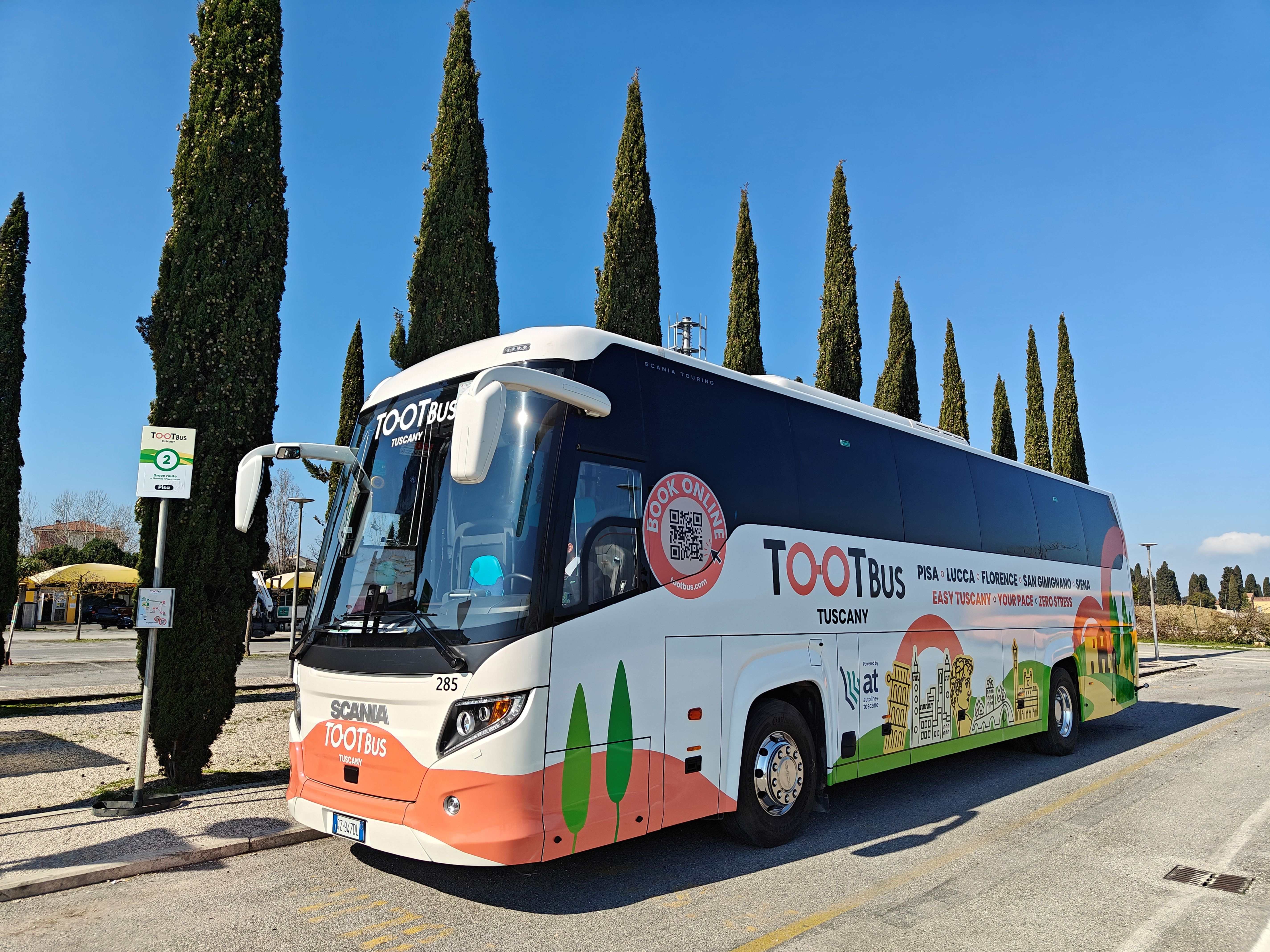 A white and orange Tootbus Tuscany coach parked on the side of a road lined with tall cypress trees under a clear blue sky.