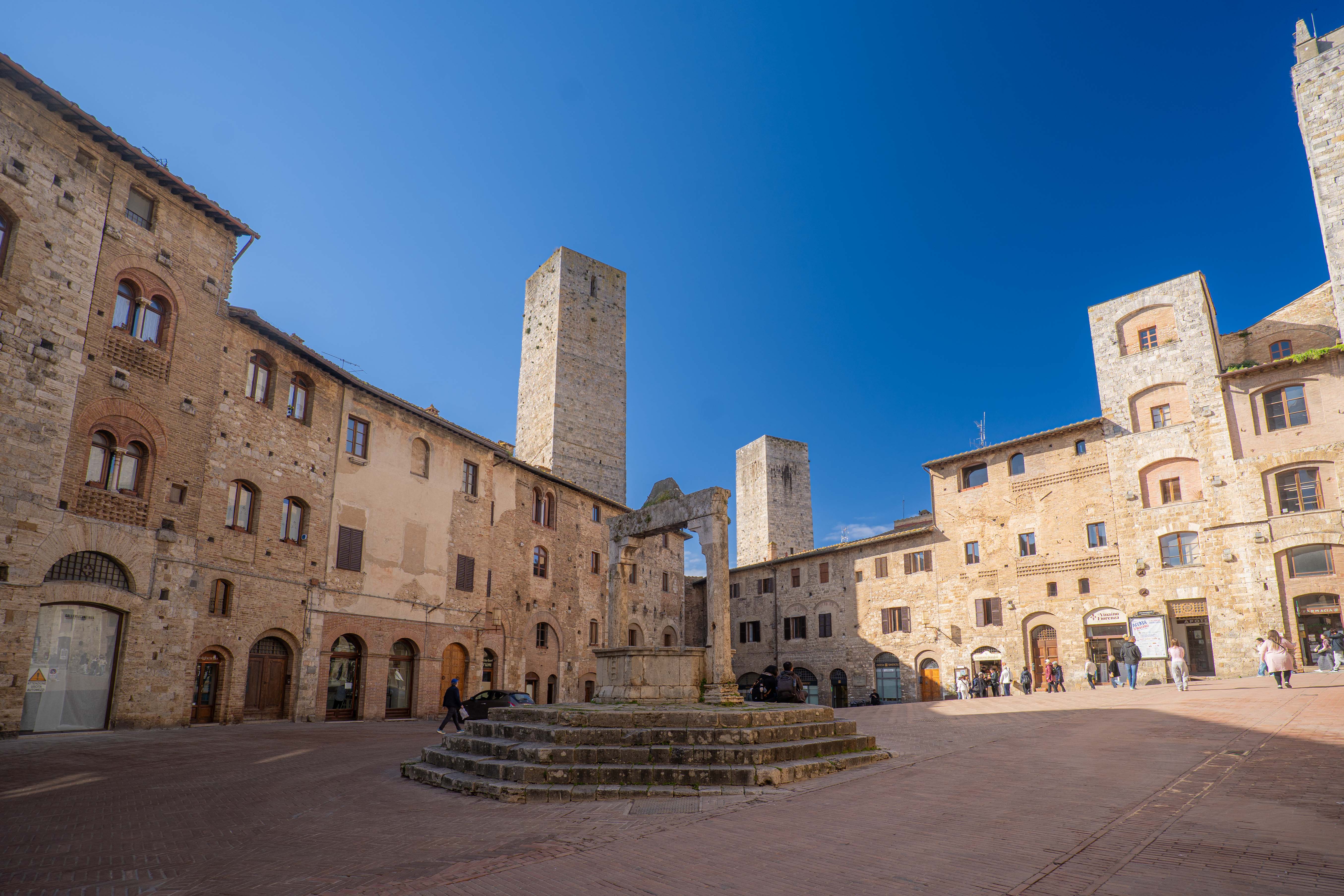 The famous Piazza della Cisterna in San Gimignano with its central stone well, surrounded by medieval houses and dominated by tall towers under a deep blue sky.