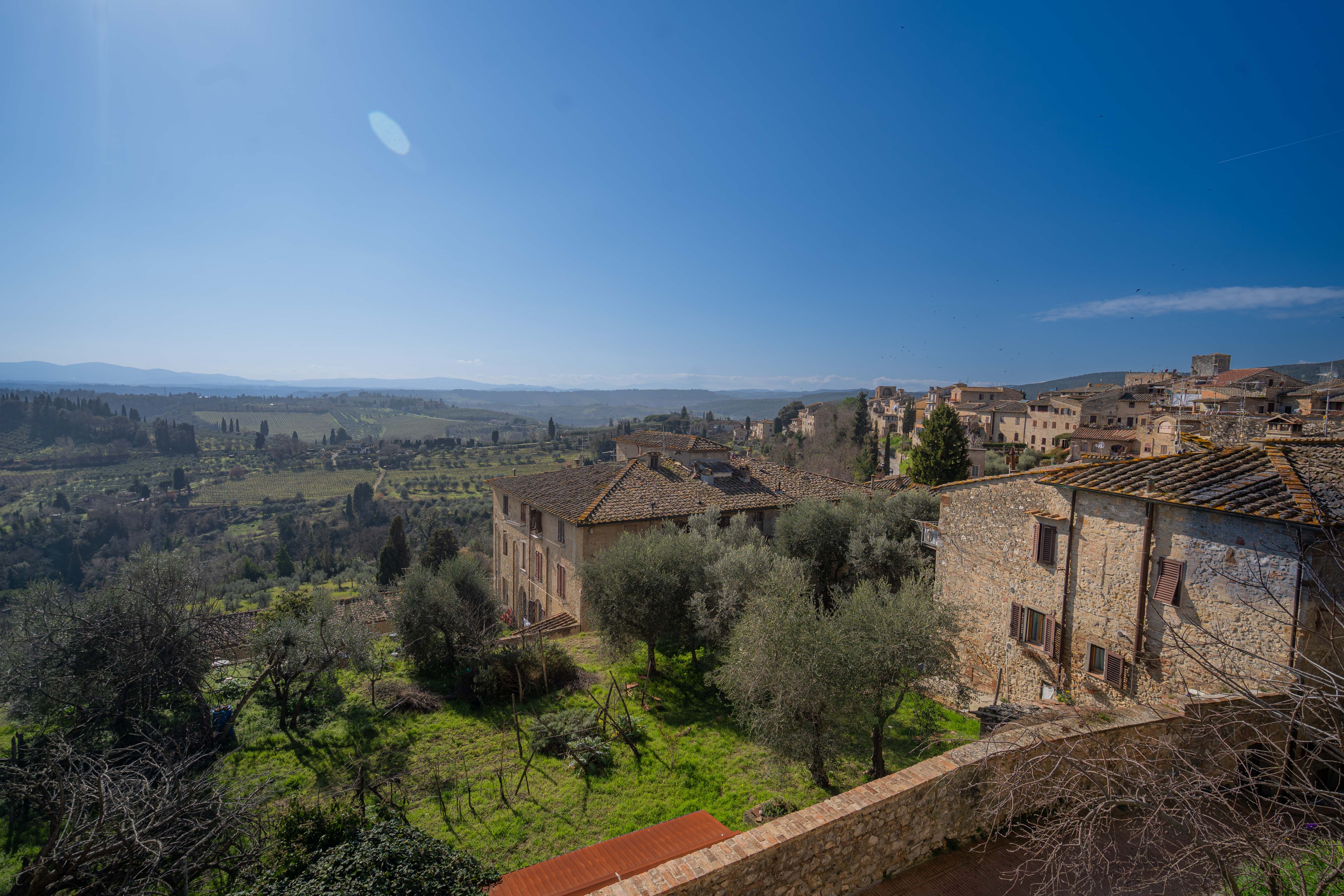 A scenic panoramic view over the tiled roofs and stone houses of San Gimignano, opening onto the green Tuscan countryside under a clear blue sky.