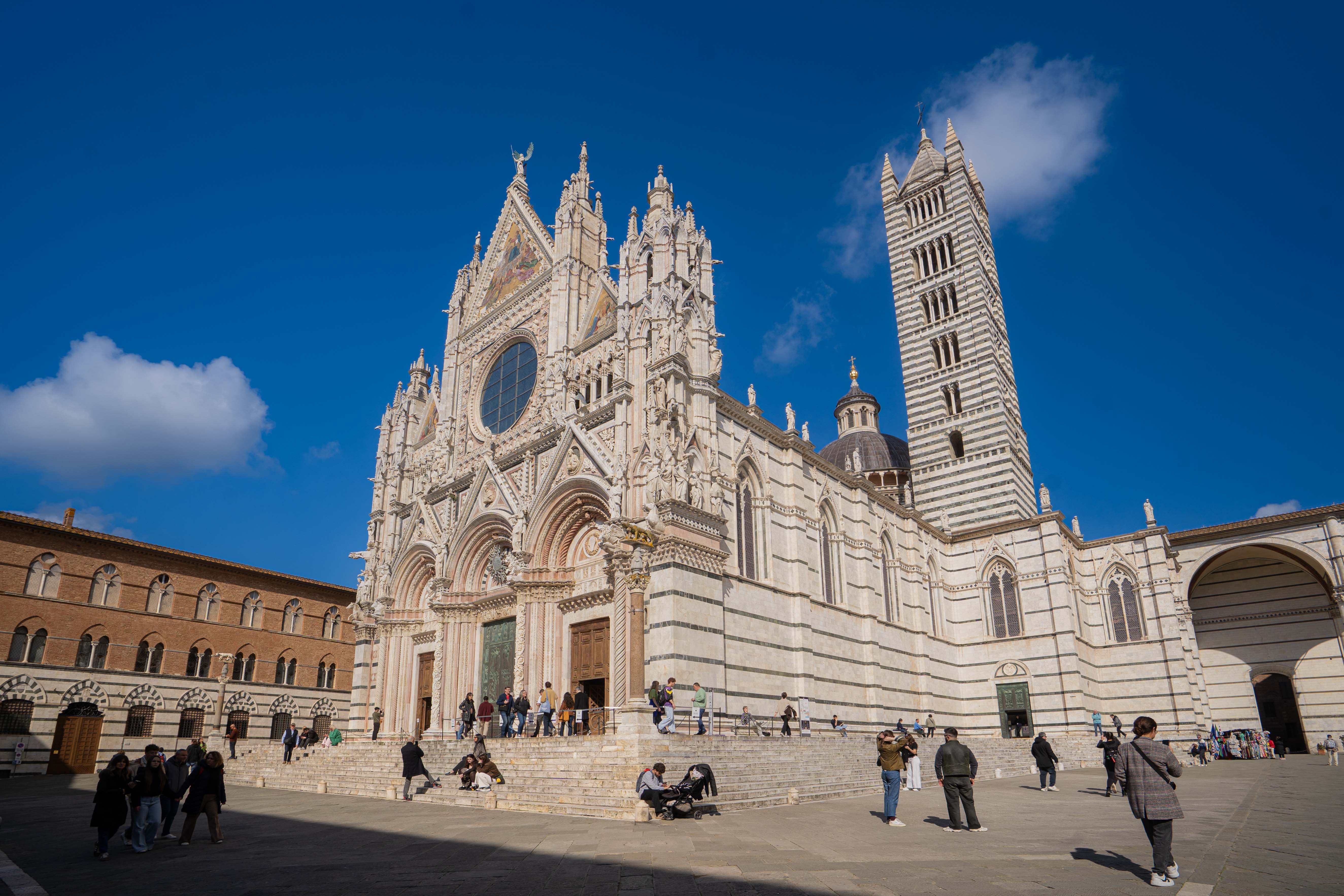 The magnificent white and black marble Gothic facade of Siena Cathedral (Duomo di Siena) rising under a blue sky with its tall campanile, one of the key stops on our new Tuscany line.