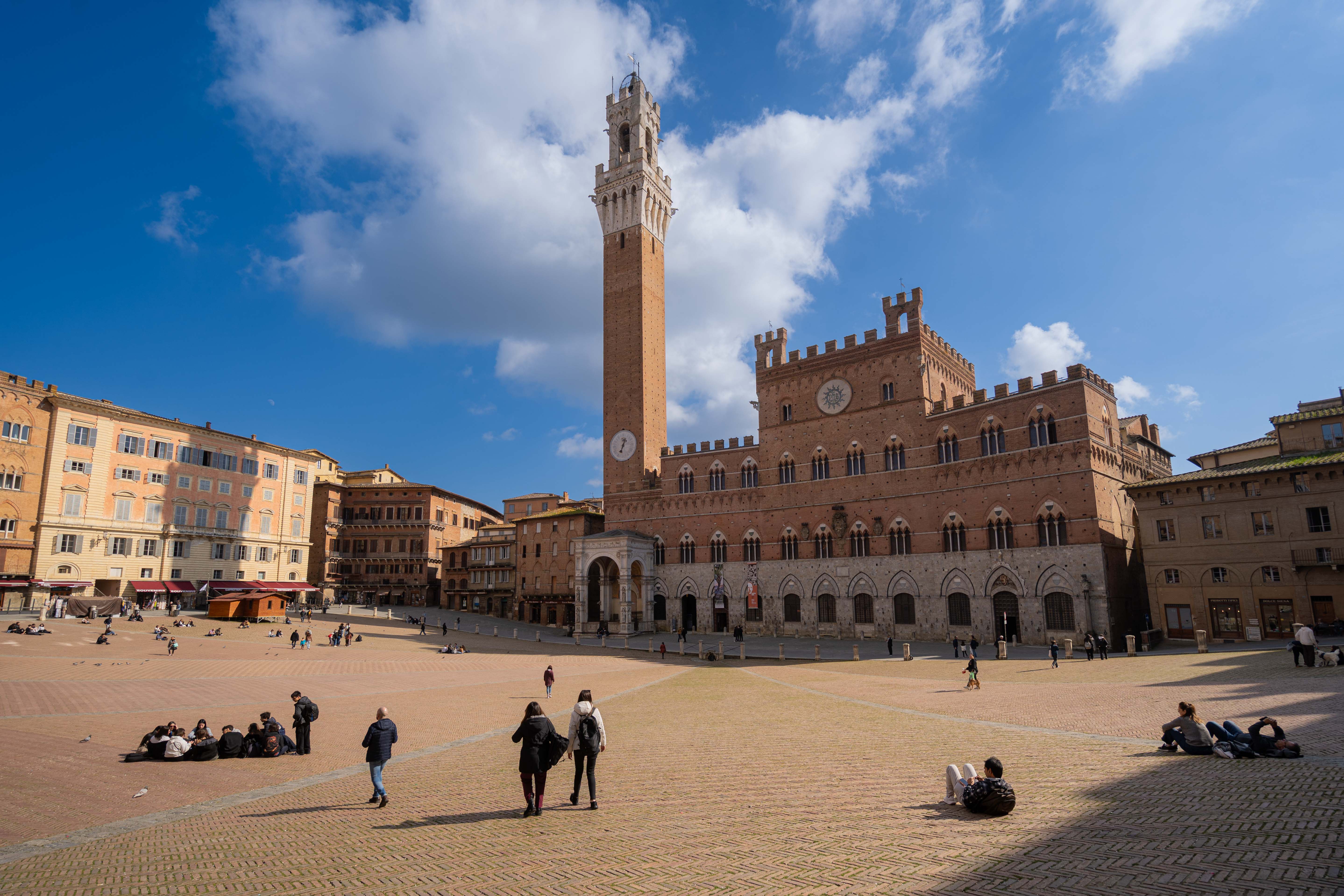 The famous Piazza del Campo in Siena under a partly cloudy blue sky, with the Palazzo Pubblico and the Torre del Mangia rising majestically, one of the highlights of our Tuscan line.