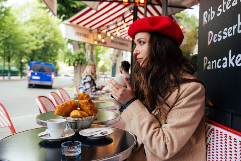 A woman with a red beret enjoys coffee and croissants on a terrace during the Emily in Paris tour.