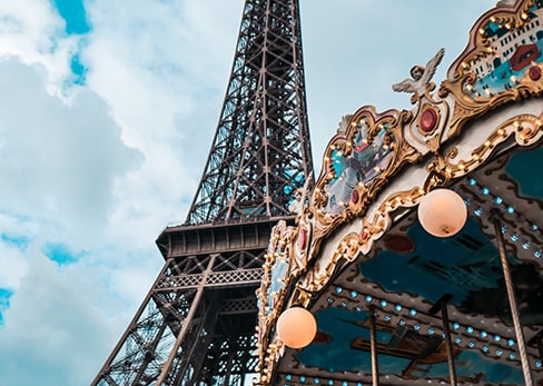 Low-angle view of the Eiffel Tower next to the illuminated details of a traditional carousel under a cloudy sky.