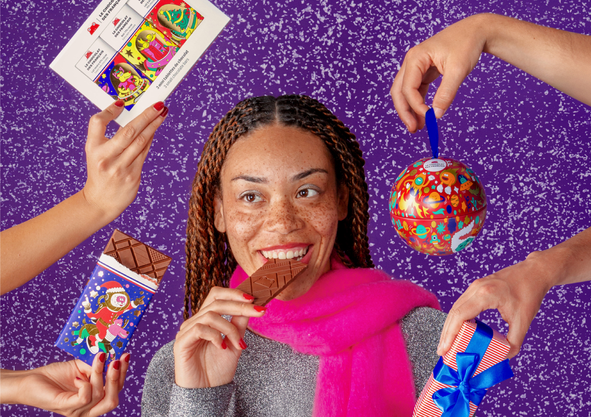 A smiling woman taking a bite of a chocolate bar, surrounded by hands presenting Le Chocolat des Français Christmas gift sets and treats against a glittery purple background.