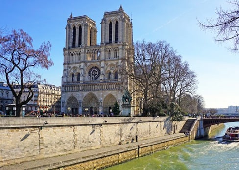 Main facade of the Notre-Dame Cathedral in Paris seen from across the Seine River on a sunny day.