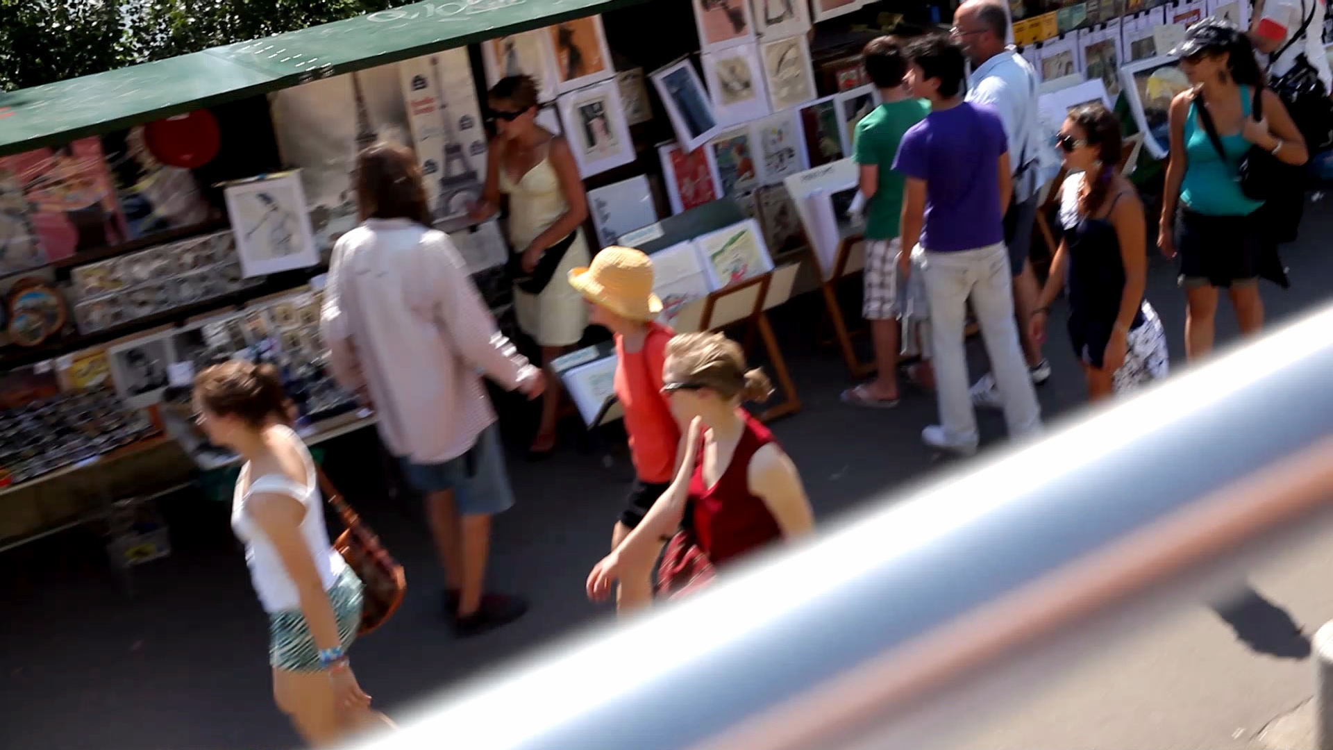 Pedestrians and tourists walk past the green stalls of booksellers displaying illustrations and souvenirs along the Seine banks.