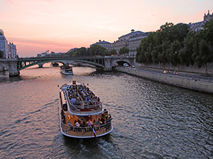 Majestic Eiffel Tower viewed from the Seine river with a cruise boat sailing in the foreground under a sunny blue sky.