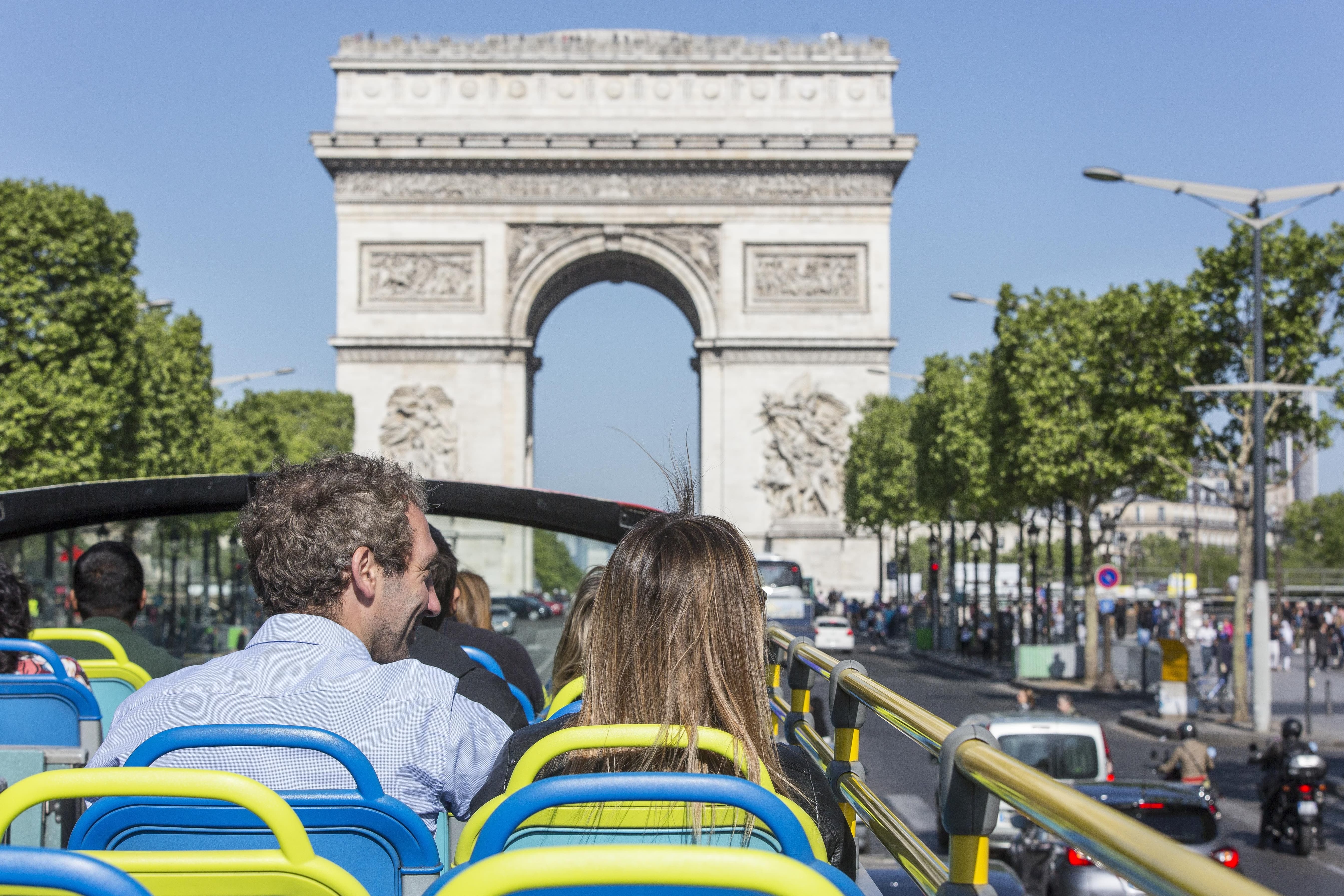 View from the open upper deck of a Tootbus tour bus, showing passengers seated from behind admiring the Arc de Triomphe standing at the end of the tree-lined Avenue des Champs-Élysées under a clear blue sky in Paris.