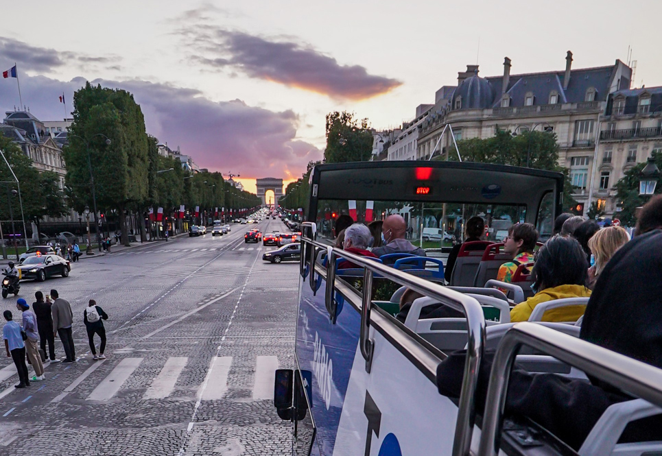 Des passagers à bord d'un bus Tootbus remontent les Champs-Élysées en direction de l'Arc de Triomphe au loin lors d'un coucher du soleil.
