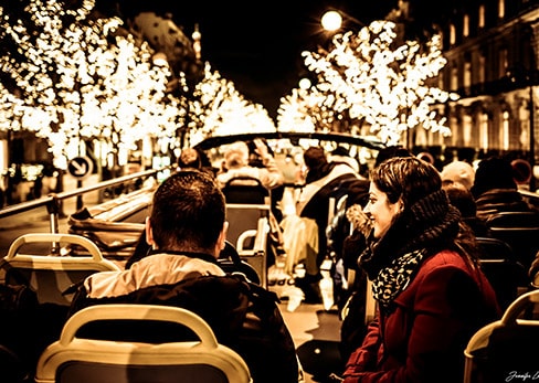 Passengers sitting on the open top deck of a Tootbus admire the Christmas lights along a Parisian avenue at night.