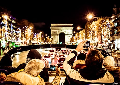 View from the open top deck of a Tootbus showing passengers from behind taking photos of the Arc de Triomphe and the Christmas lights along the Champs-Élysées avenue at night.