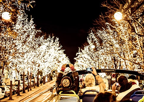 Passengers on the top deck of a Tootbus take photos with their smartphones of illuminated trees along a Parisian avenue at night.