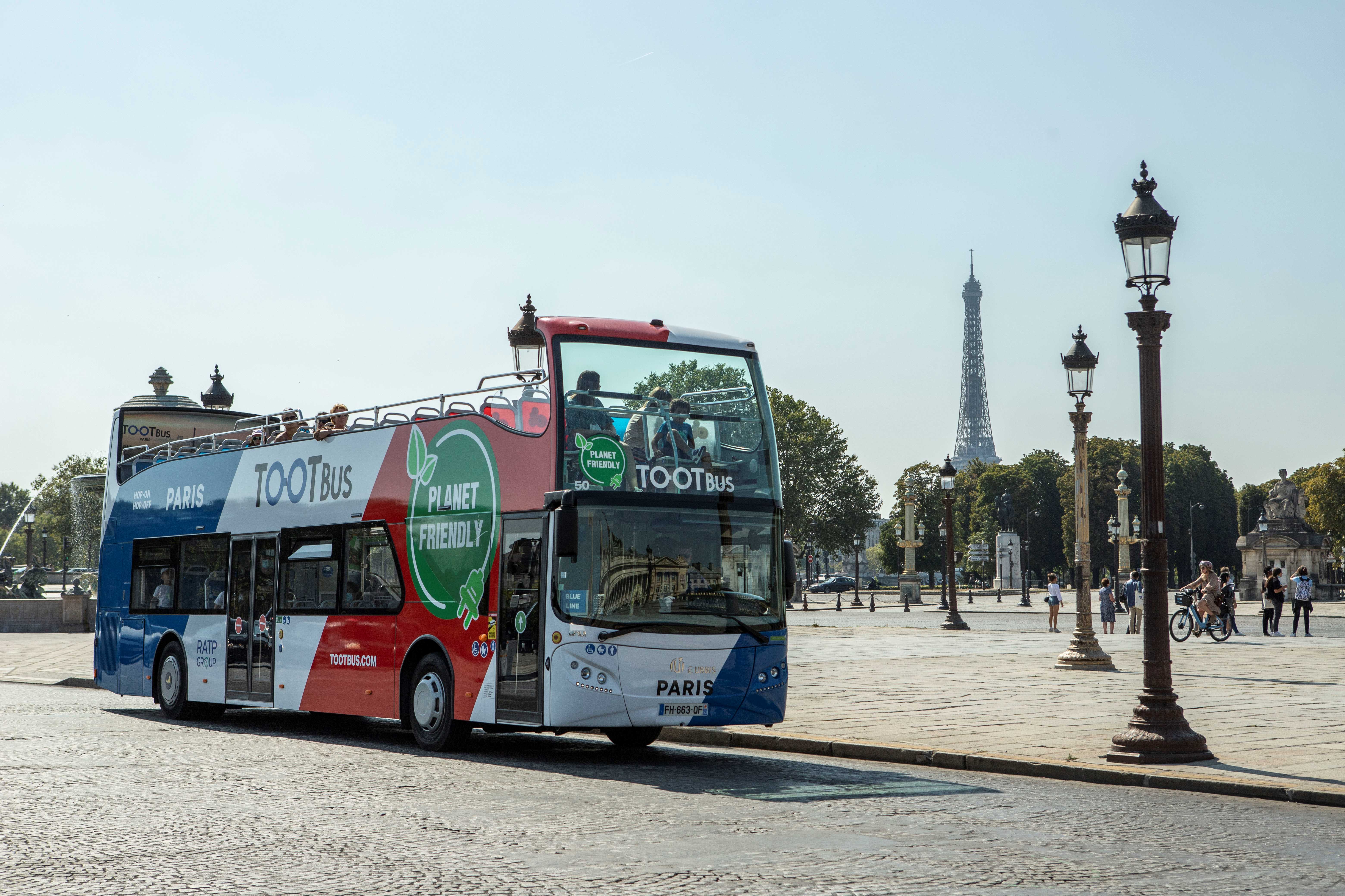 Vue de trois-quarts avant d'un bus touristique à impériale Tootbus bleu, blanc et rouge, circulant sur les pavés de la place de la Concorde à Paris, avec des lampadaires historiques au premier plan et la Tour Eiffel visible au loin sous un ciel clair.