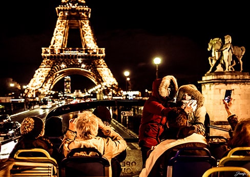 Passengers on board the open top deck of a Tootbus take photos and selfies in front of the illuminated Eiffel Tower at night.