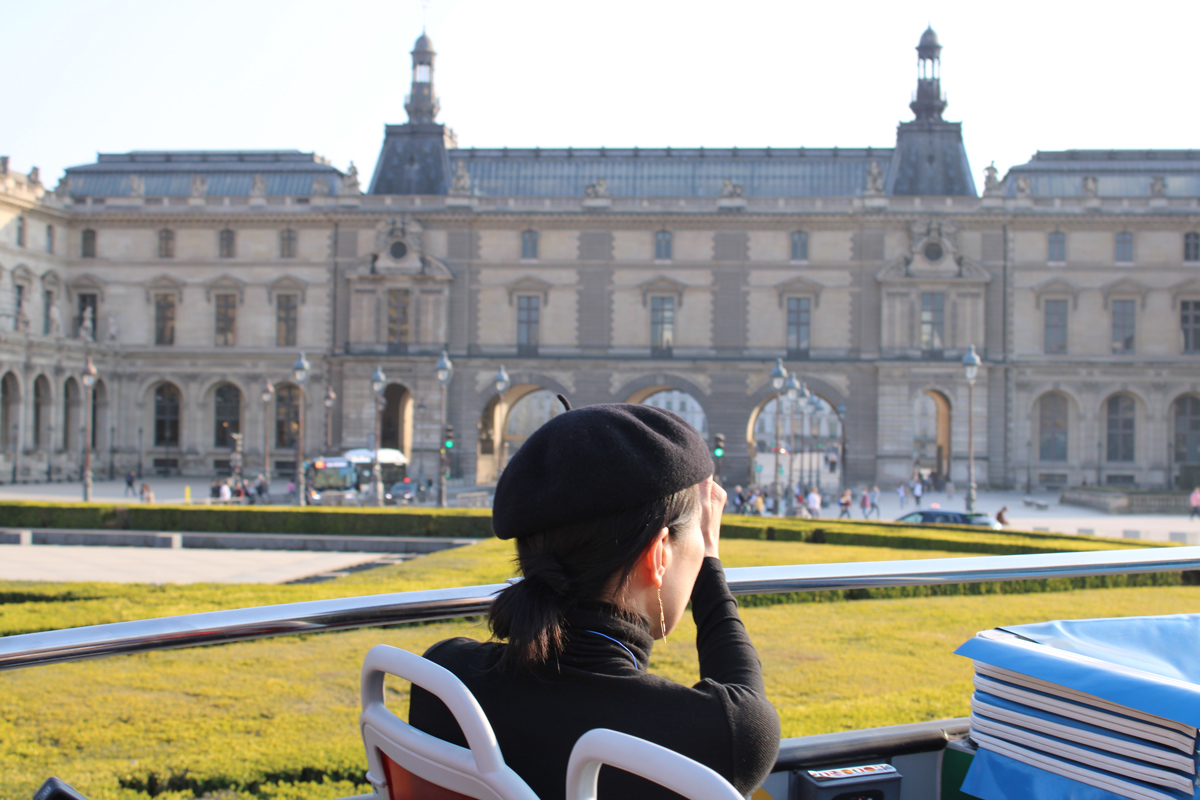 Une passagère portant un béret noir vue de dos à bord d'un bus Tootbus regarde le musée du Louvre à Paris.