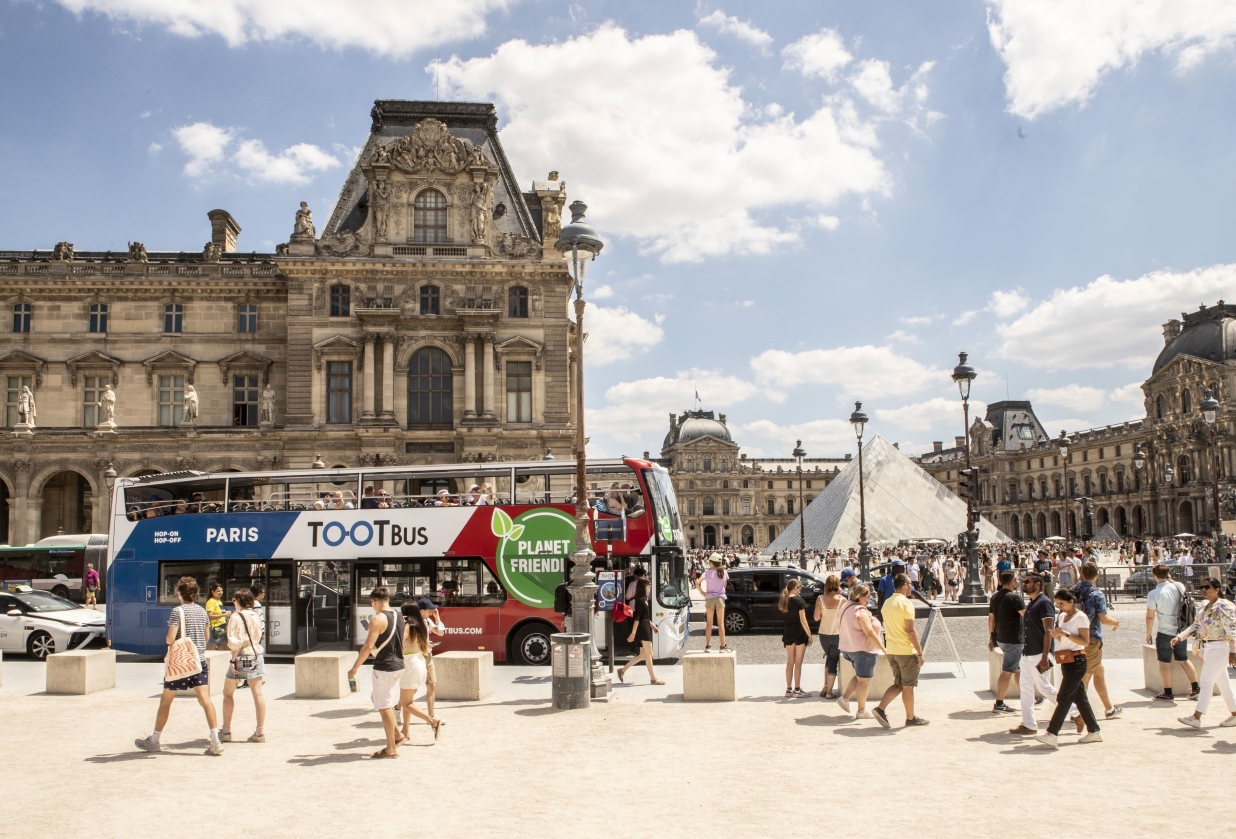 A Tootbus double-decker bus parked at the Louvre Museum square with pedestrians in the foreground and the glass pyramid in the background.