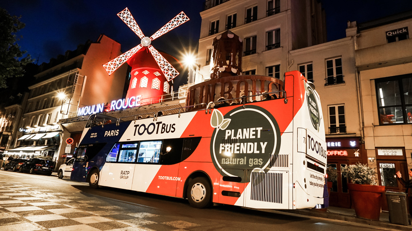 Un bus écoresponsable Tootbus fonctionnant au gaz naturel passe devant le célèbre Moulin Rouge illuminé la nuit à Paris.