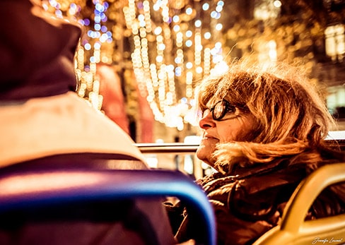 A smiling woman with glasses sitting on the top deck of a Tootbus admires the night lights in Paris.