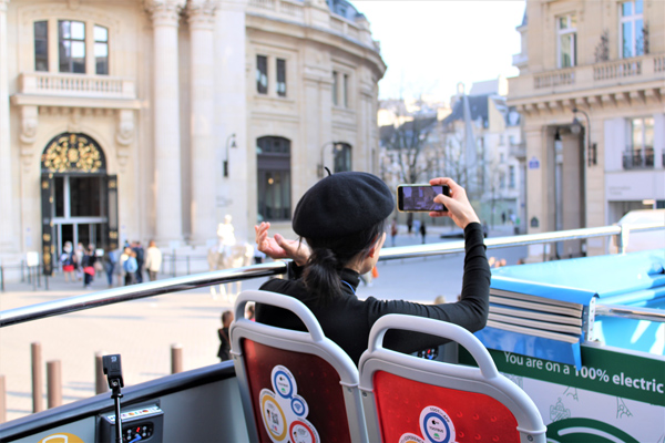 Une passagère portant un béret noir prend une photo avec son smartphone depuis l'étage ouvert d'un bus électrique Tootbus devant l'Opéra Garnier.