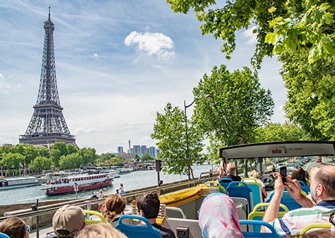 View from the open upper deck of a Tootbus tour bus, showing passengers seated from behind admiring the Arc de Triomphe standing at the end of the tree-lined Avenue des Champs-Élysées under a clear blue sky in Paris.