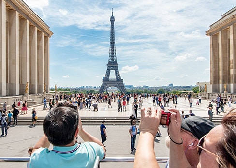 View from the open upper deck of a Tootbus tour bus, showing passengers in the foreground taking photos with their phones and cameras of the Eiffel Tower rising in the center of the Trocadéro esplanade bustling with visitors under a clear sky.