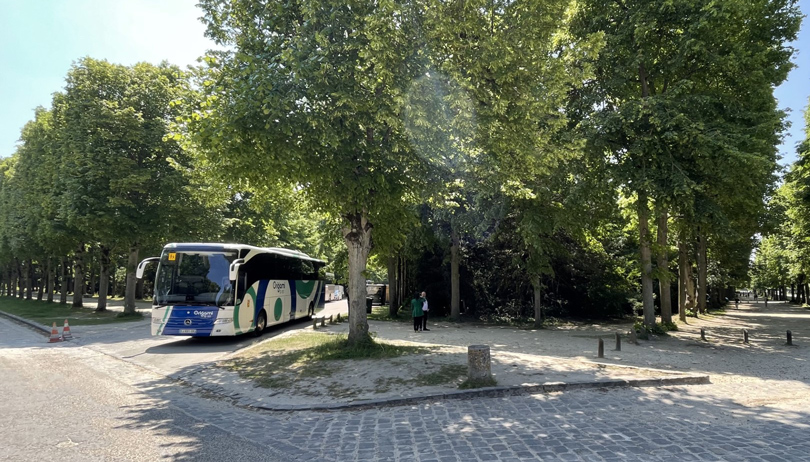 Un autocar de Tootbus estacionado junto a un paseo arbolado y sombreado cerca de los jardines de Versalles en un día soleado.