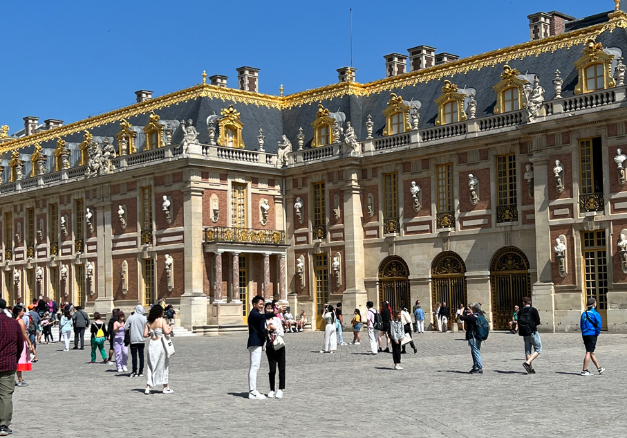 Des touristes se promènent et prennent des photos dans la Cour de Marbre du Château de Versailles devant les façades ornées de bustes et de dorures.