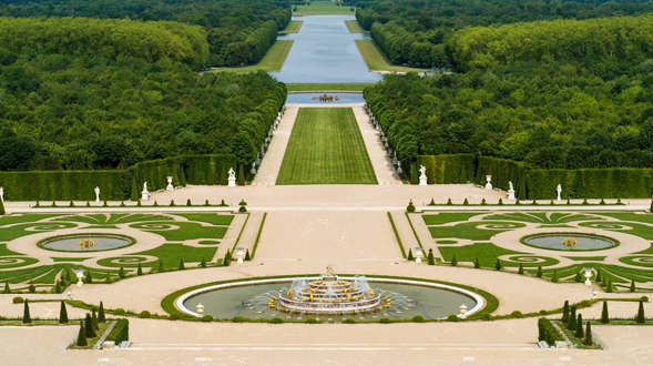 Una vista elevada y en perspectiva de los jardines de Versalles, que destaca la Fuente de Latona, el Tapis Vert y el Gran Canal extendiéndose hacia el horizonte.